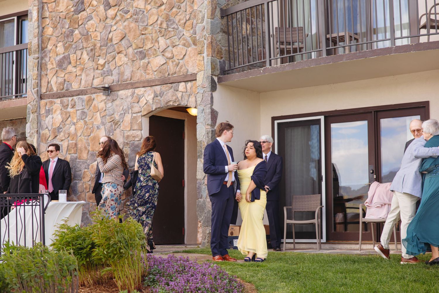 Wedding guests mingling outside a rustic stone venue building on a sunny afternoon.