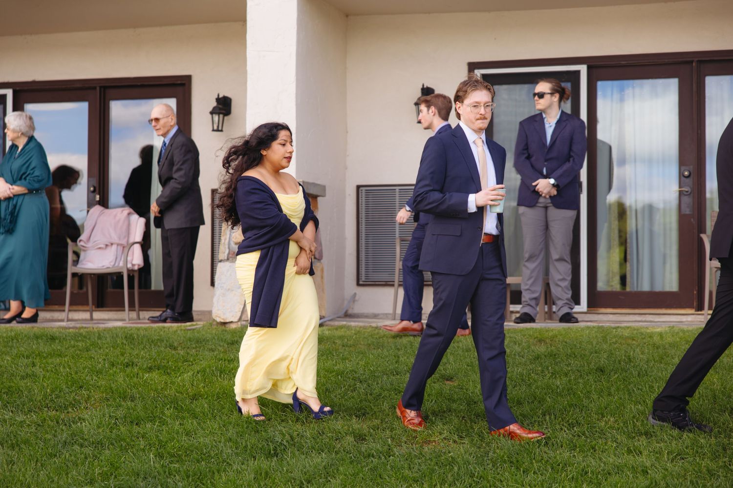 Wedding guests in formal attire walking on green lawn outside elegant venue during outdoor reception.
