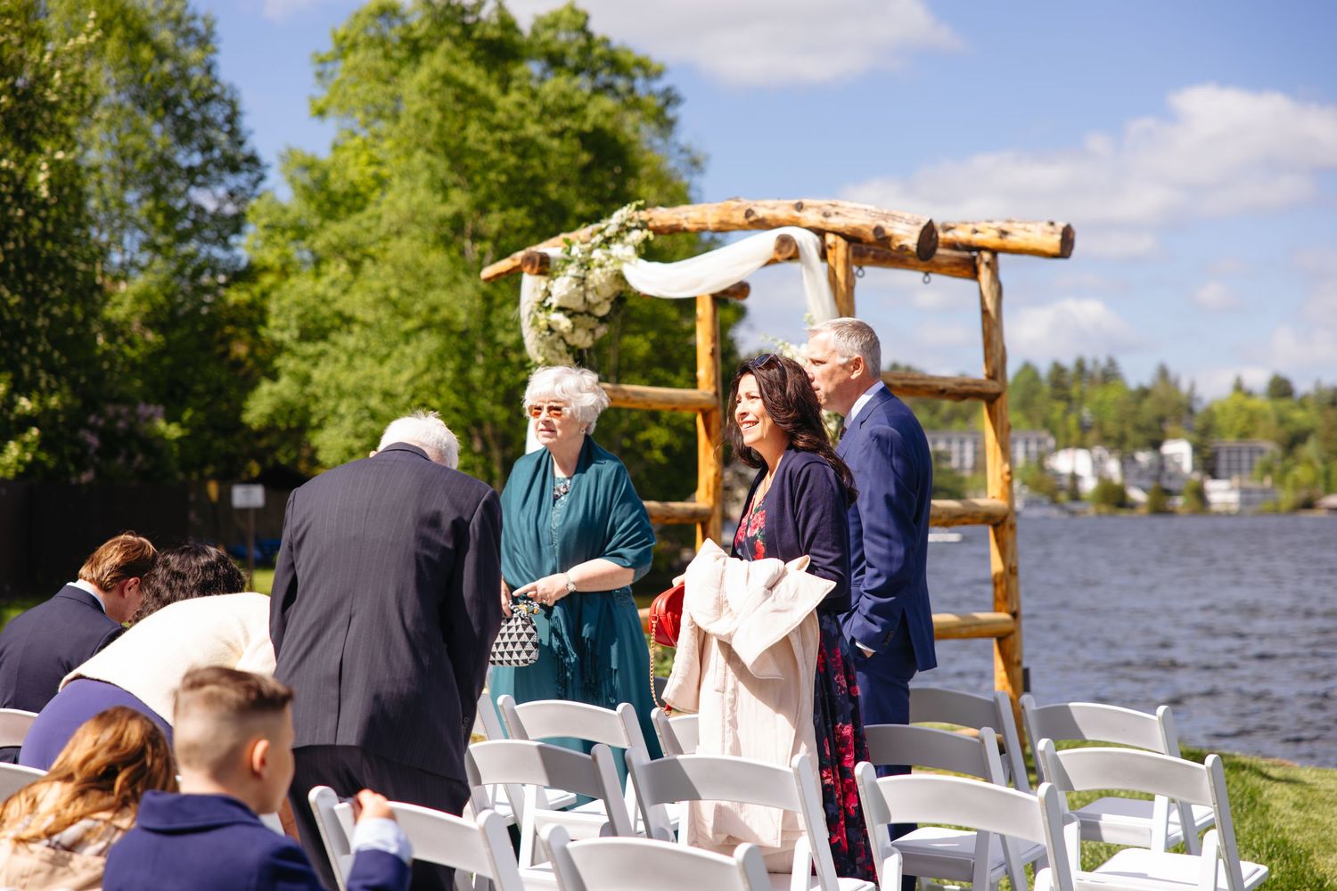 Wedding guests gathering near rustic wooden arch beside a lake on a sunny day before the ceremony.