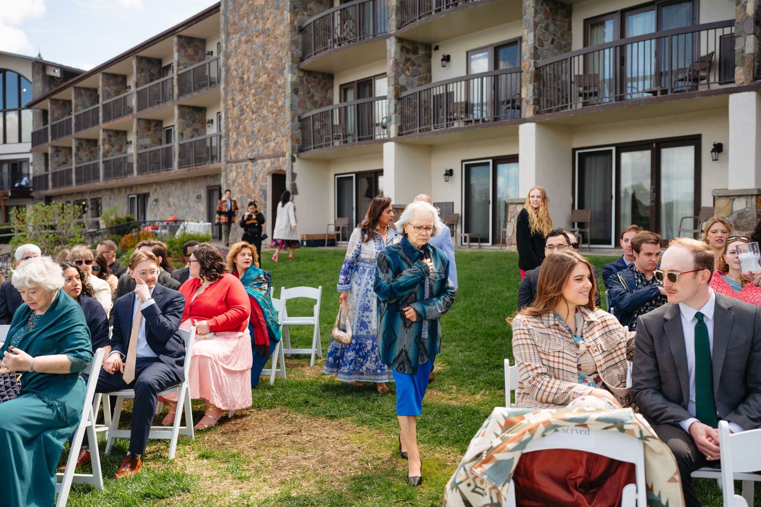 Guests gather on a lawn for an outdoor wedding ceremony beside a multi-story hotel building.