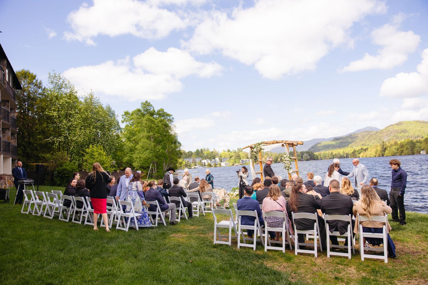 Wedding guests seated in white chairs lakeside under blue skies for an outdoor ceremony.