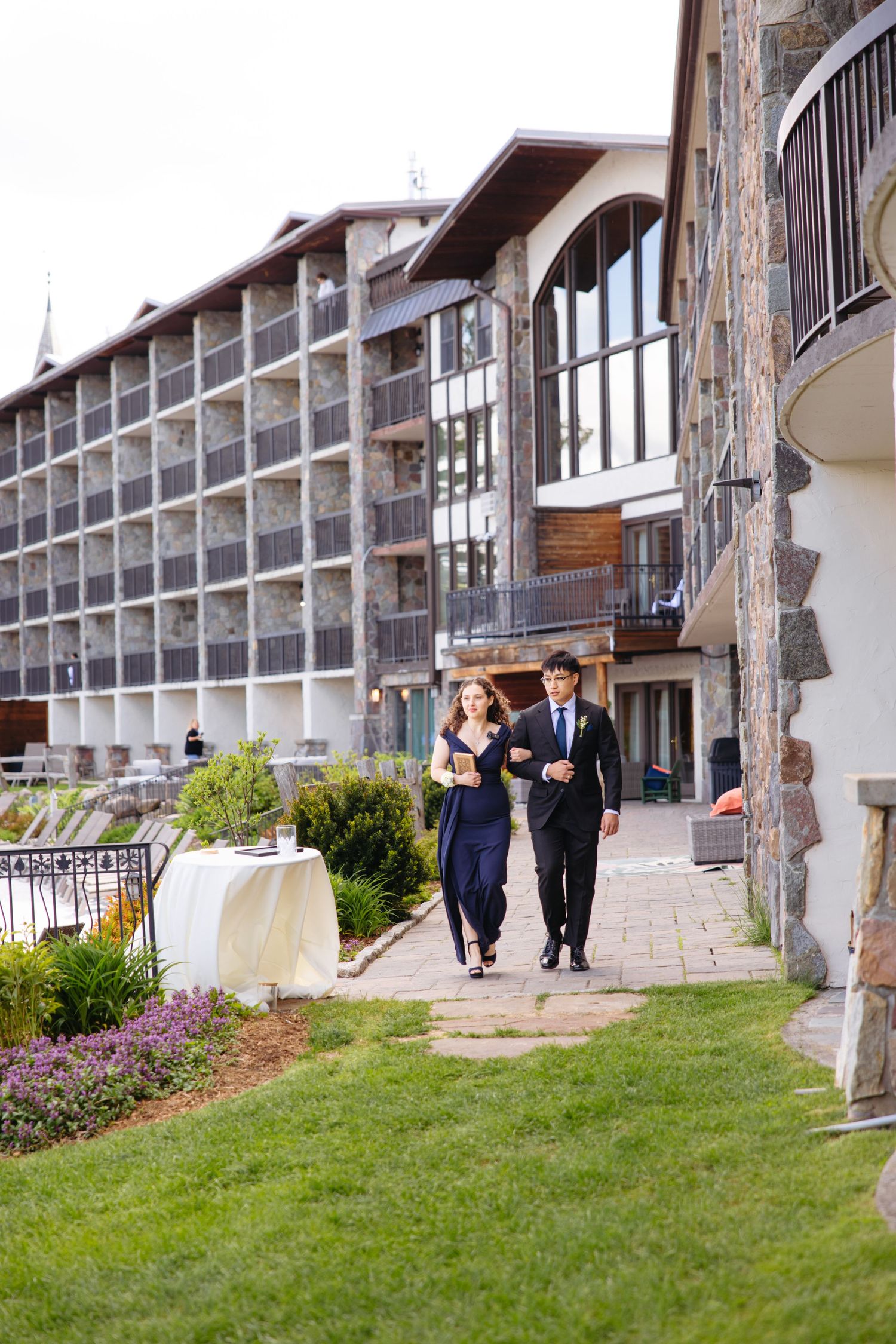 Elegant couple in formal attire walking along a stone path outside a luxury resort hotel.