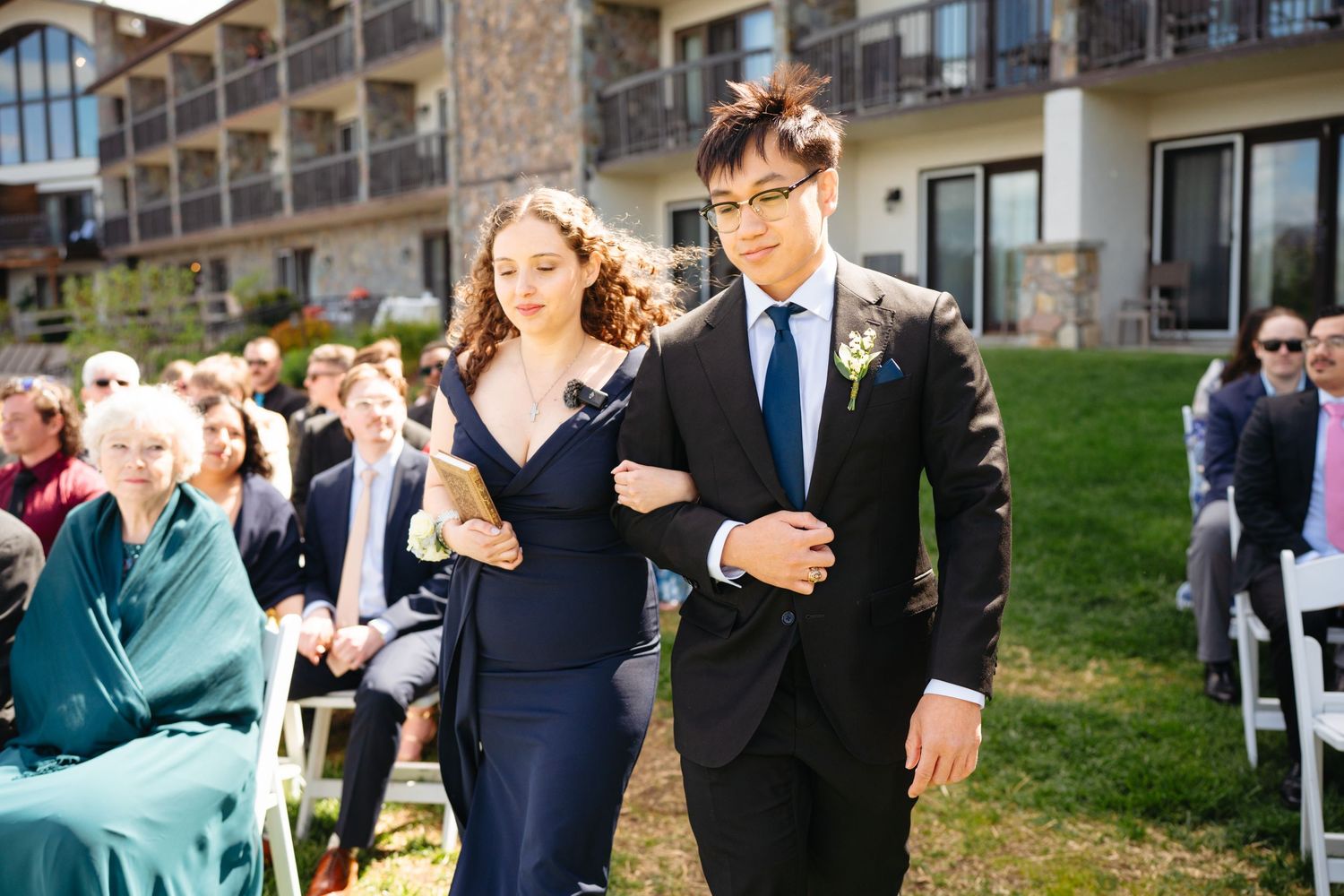 Bridesmaid and groomsman walking down outdoor wedding aisle at lakeside resort on sunny spring day.