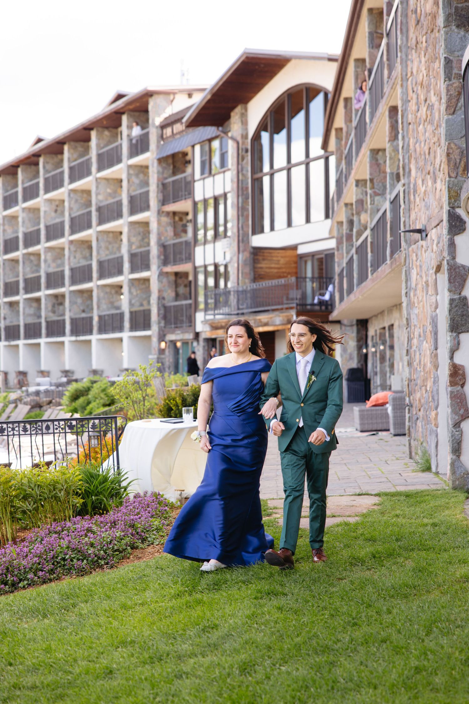 Couple in elegant formalwear, blue gown and green suit, posing outside a luxury stone hotel on manicured grounds.