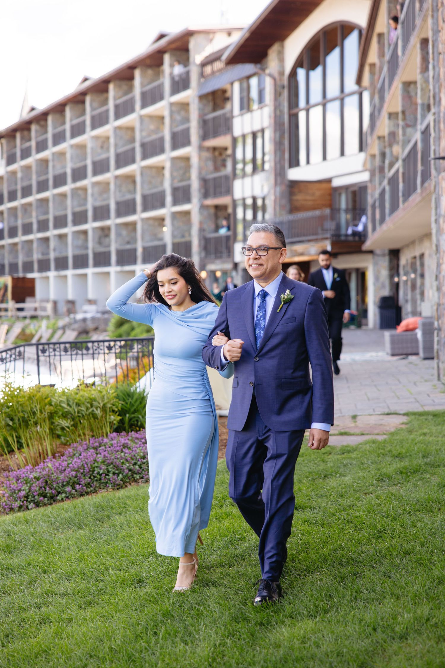 Woman in light blue dress and man in navy suit walking arm-in-arm outside modern lakeside wedding venue.