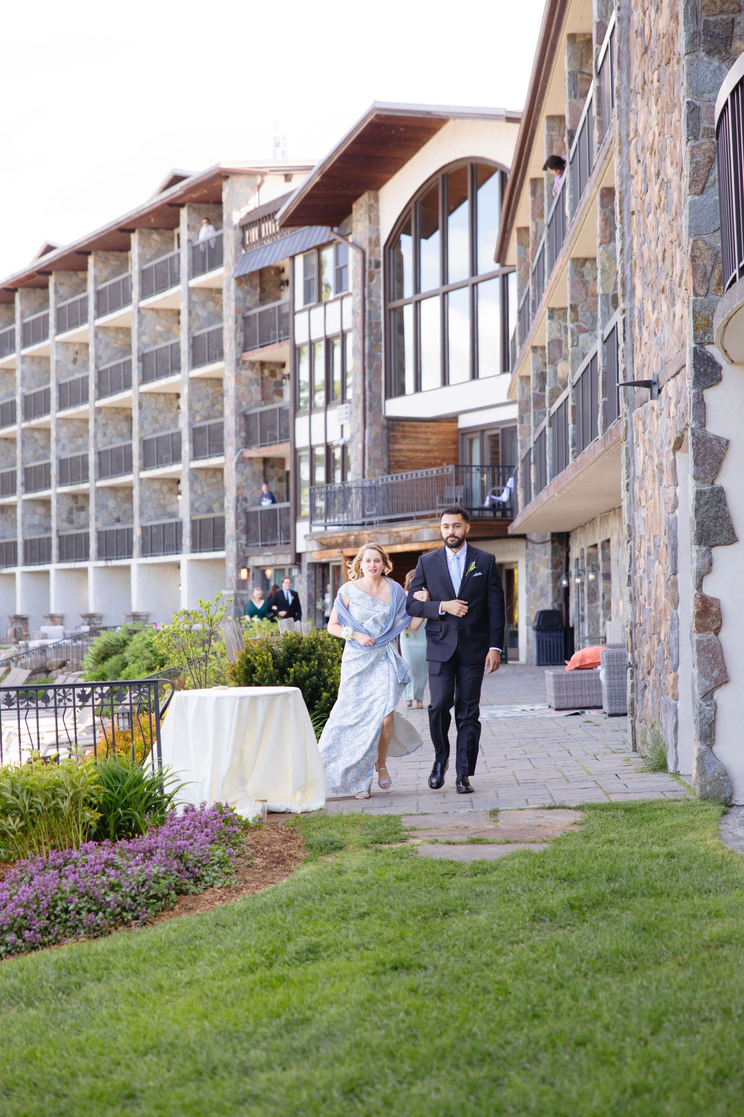 Couple walking along stone pathway beside a lakeside resort hotel on a wedding day.