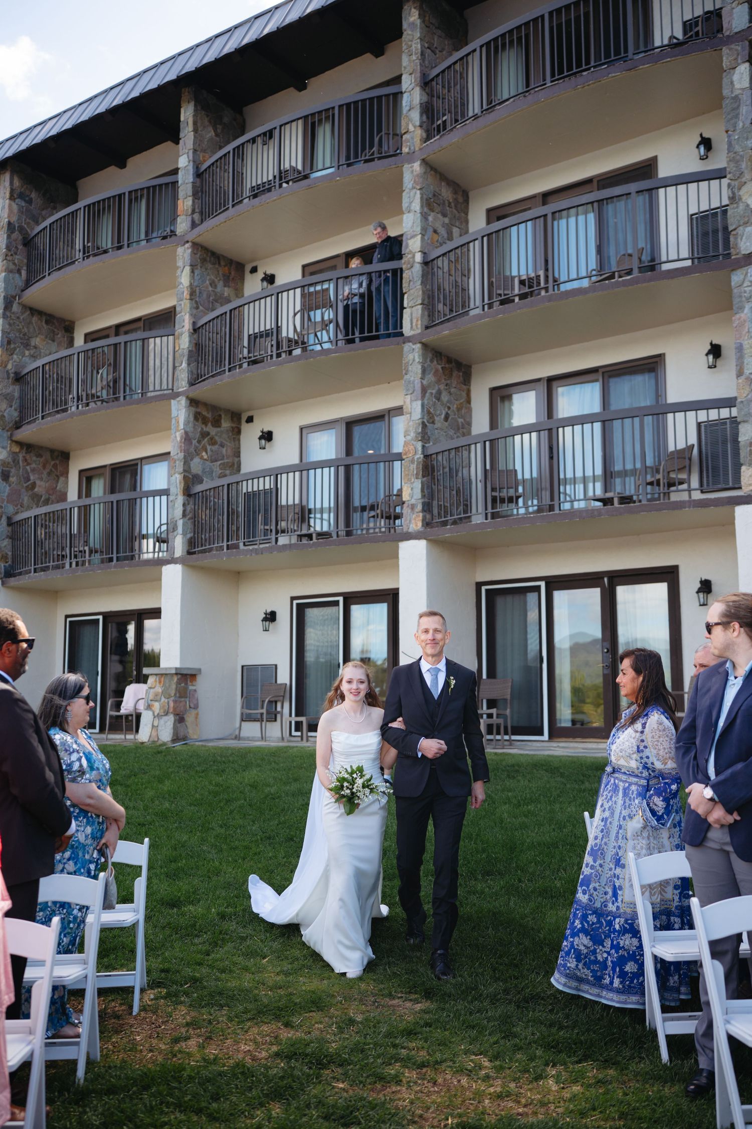 Bride and groom walking down outdoor aisle at wedding ceremony with stone hotel balconies in background.