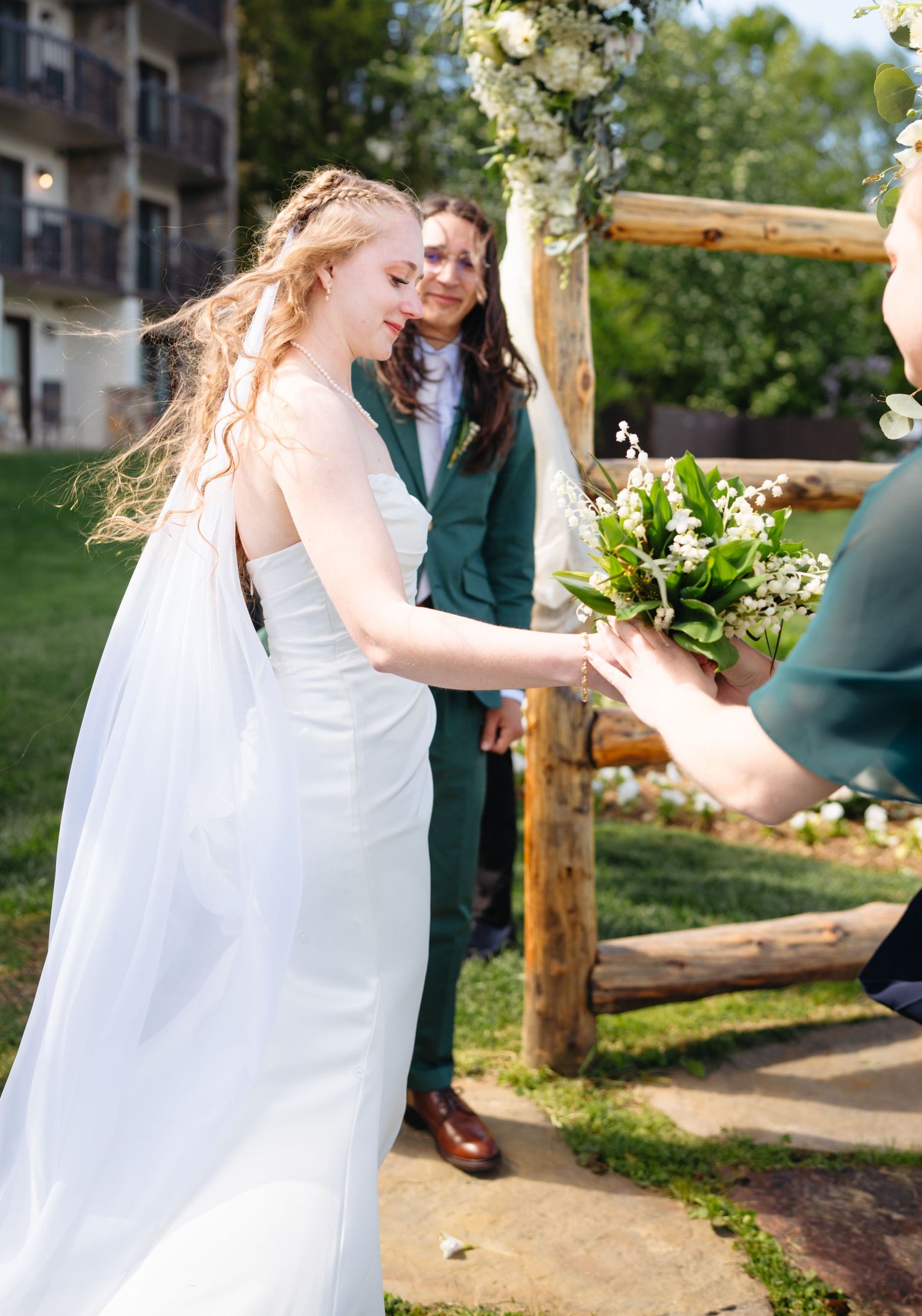 Bride in white gown receiving lily of the valley bouquet at outdoor rustic log arch ceremony.