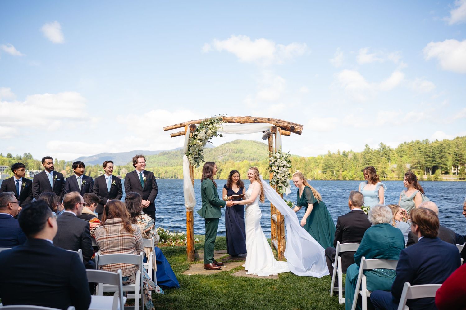 Outdoor lakeside wedding ceremony with couple at rustic wooden floral arch, groomsmen, and seated guests.