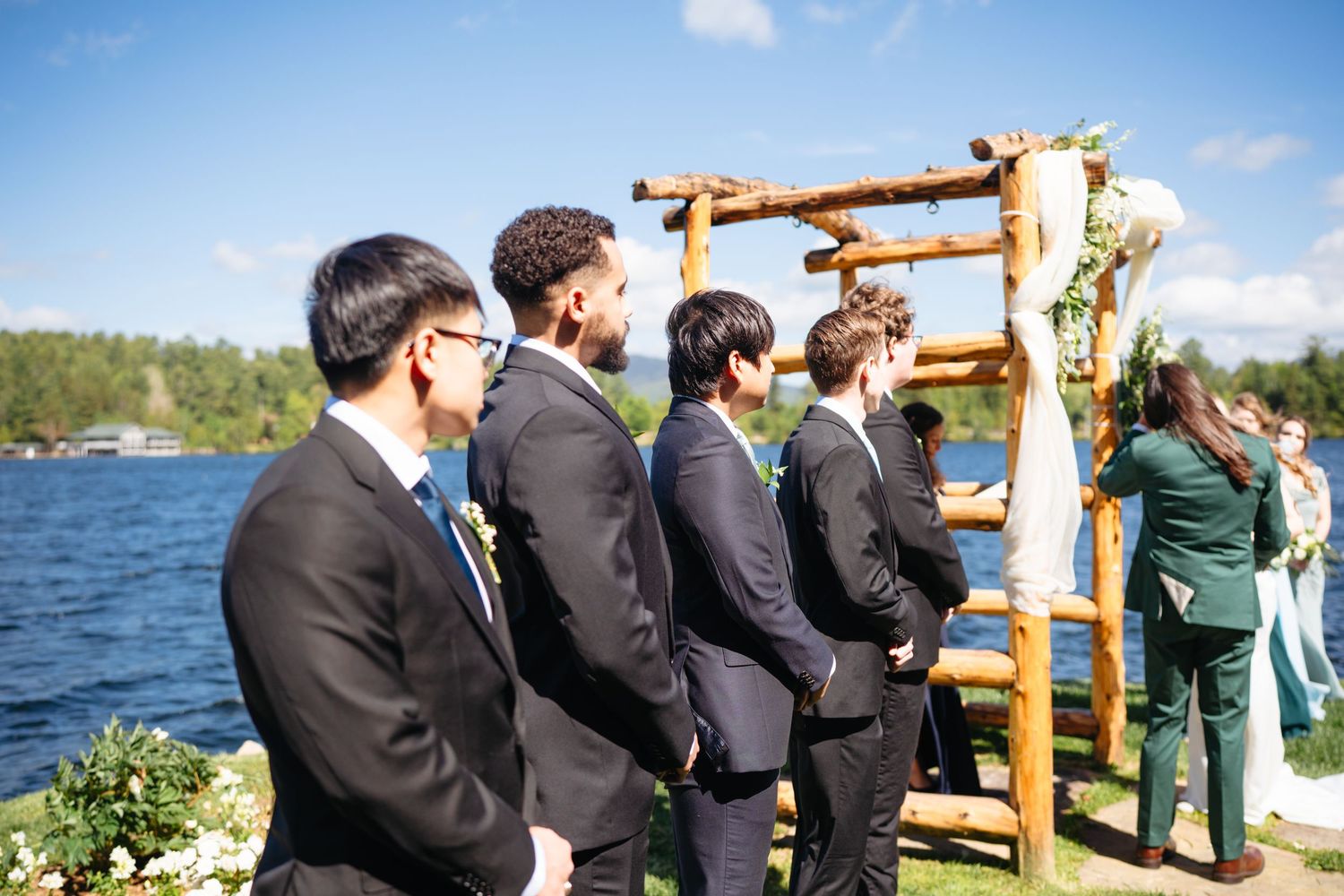 Groomsmen in dark suits standing at an outdoor lakeside wedding ceremony with wooden arch.