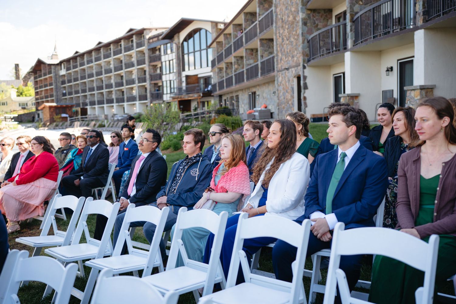 Wedding guests seated in white chairs at an outdoor ceremony beside a multi-story stone resort hotel.