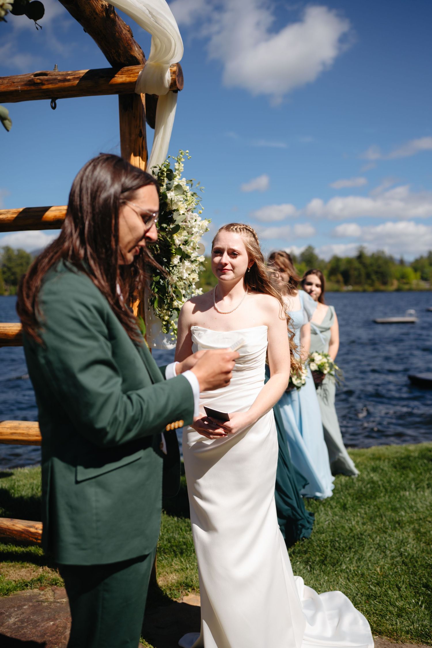 Bride in white gown smiling at officiant during outdoor lakeside wedding ceremony.