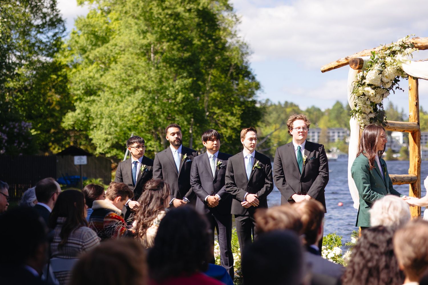 Five groomsmen in dark suits standing at attention beside floral arch during outdoor lakeside ceremony.