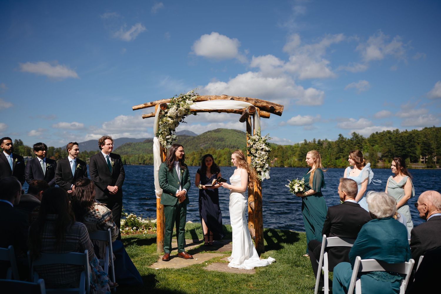 Bride and groom exchanging vows under rustic wooden arch at scenic lakeside outdoor wedding ceremony.