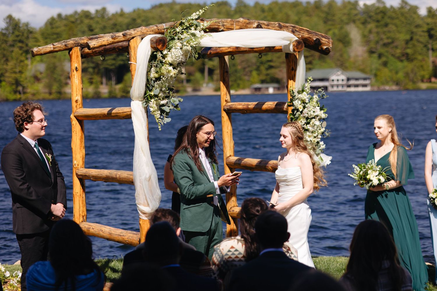 Couple exchanges vows under a rustic wooden arch adorned with white flowers beside a serene lake.
