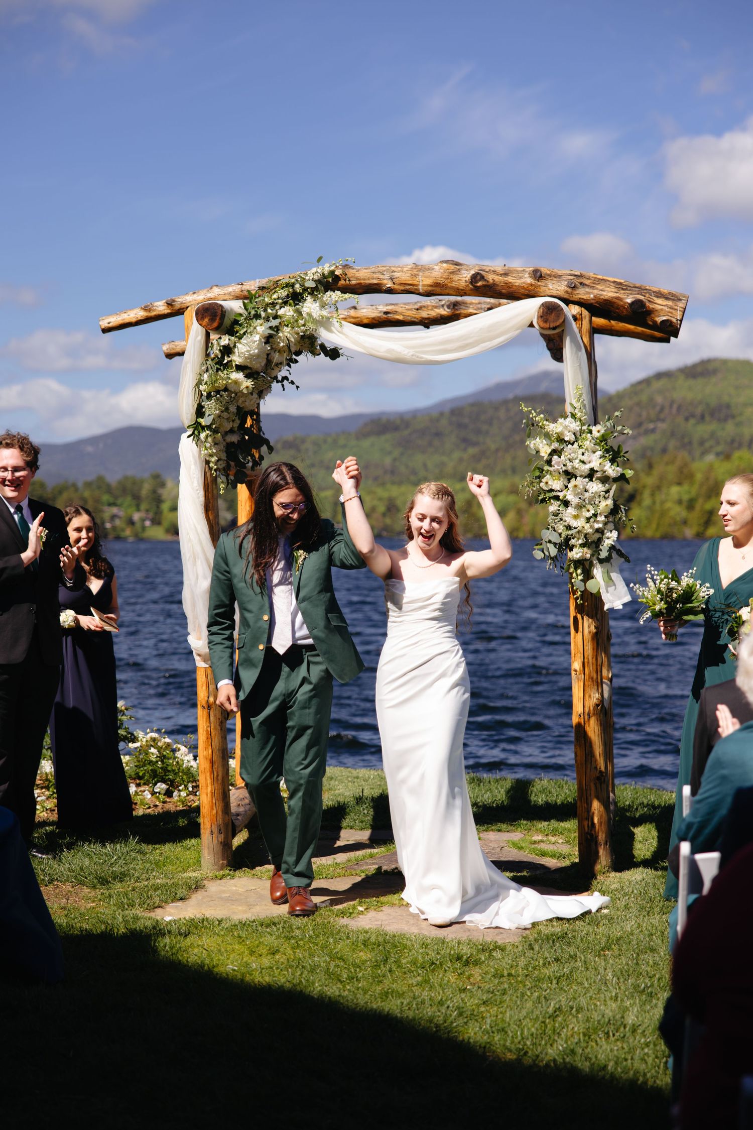 Joyful bride raises arms under rustic wooden arch with flowers by a lake as groom walks beside her.