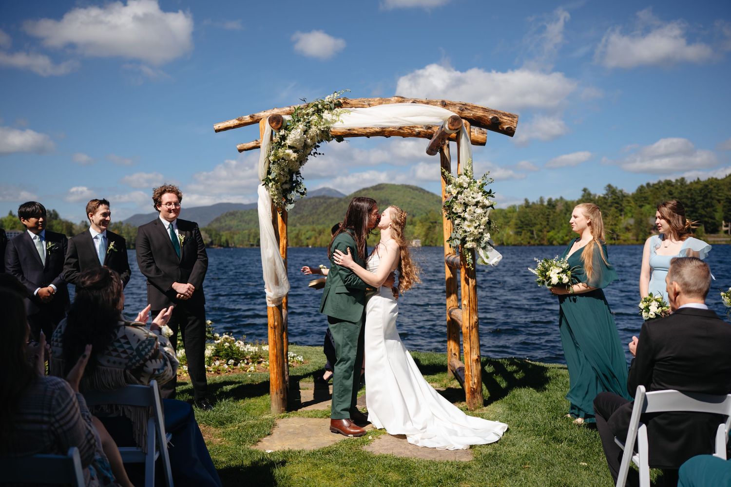 Bride and groom exchanging vows under rustic log arch by lakeside with mountain backdrop on sunny day.
