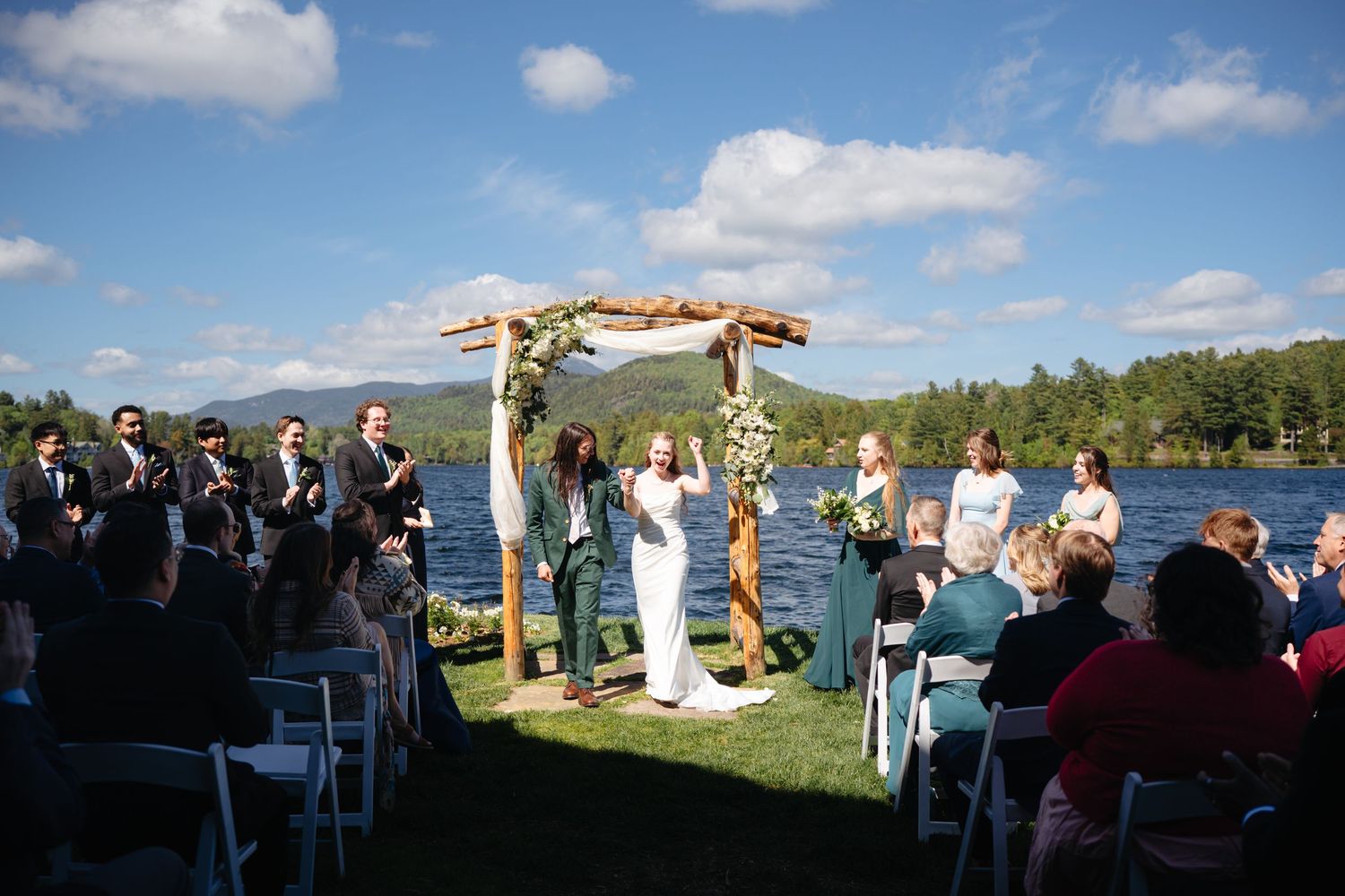 Outdoor lakeside wedding ceremony with couple at rustic wooden arch, guests seated in white chairs.