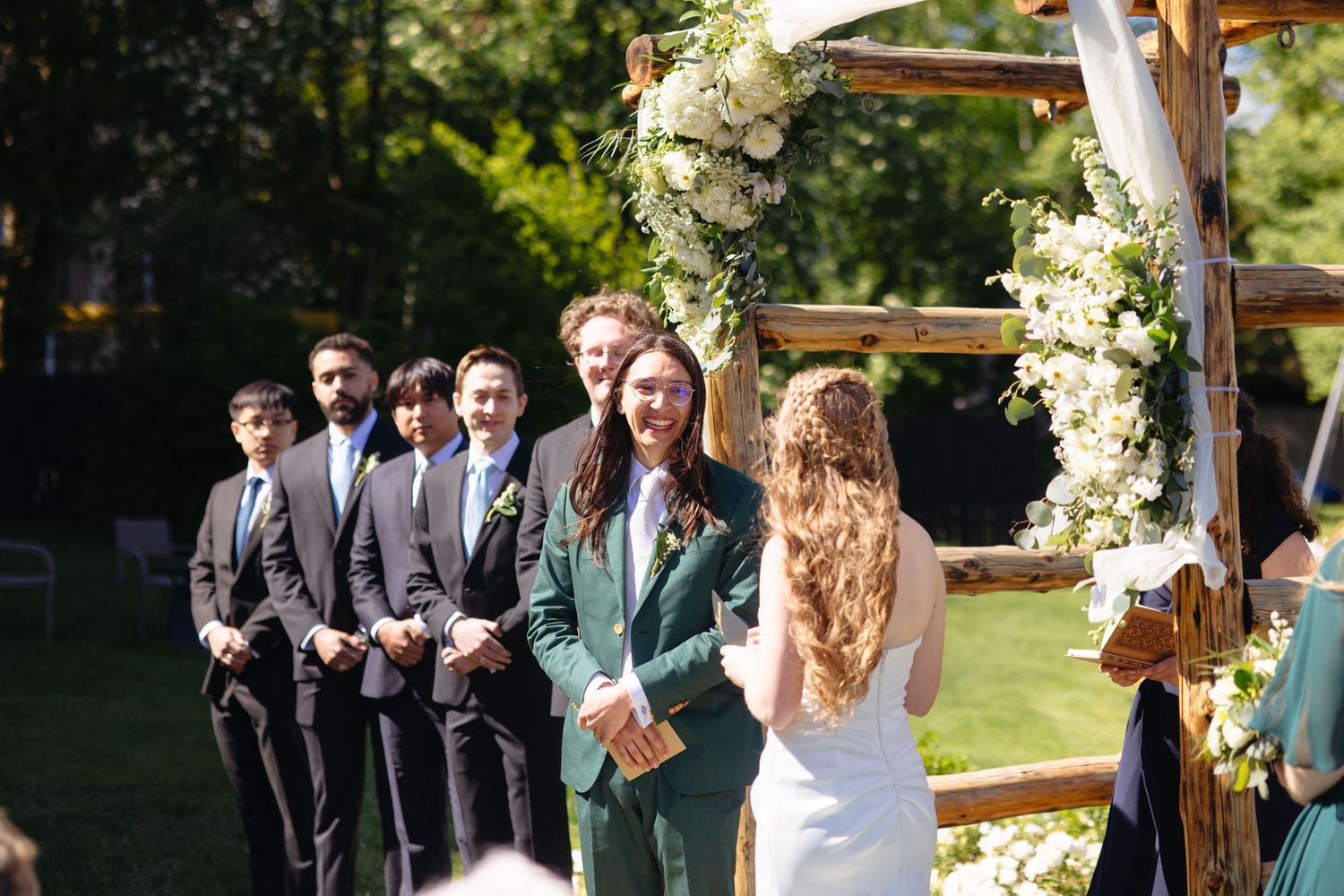 Groom in green suit smiling at bride at outdoor wedding ceremony with floral arch.