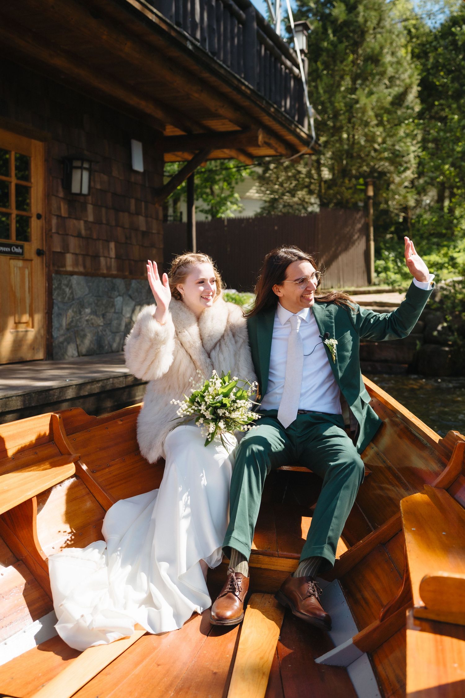Happy bride and groom waving while seated together in a wooden boat outside rustic waterfront building.