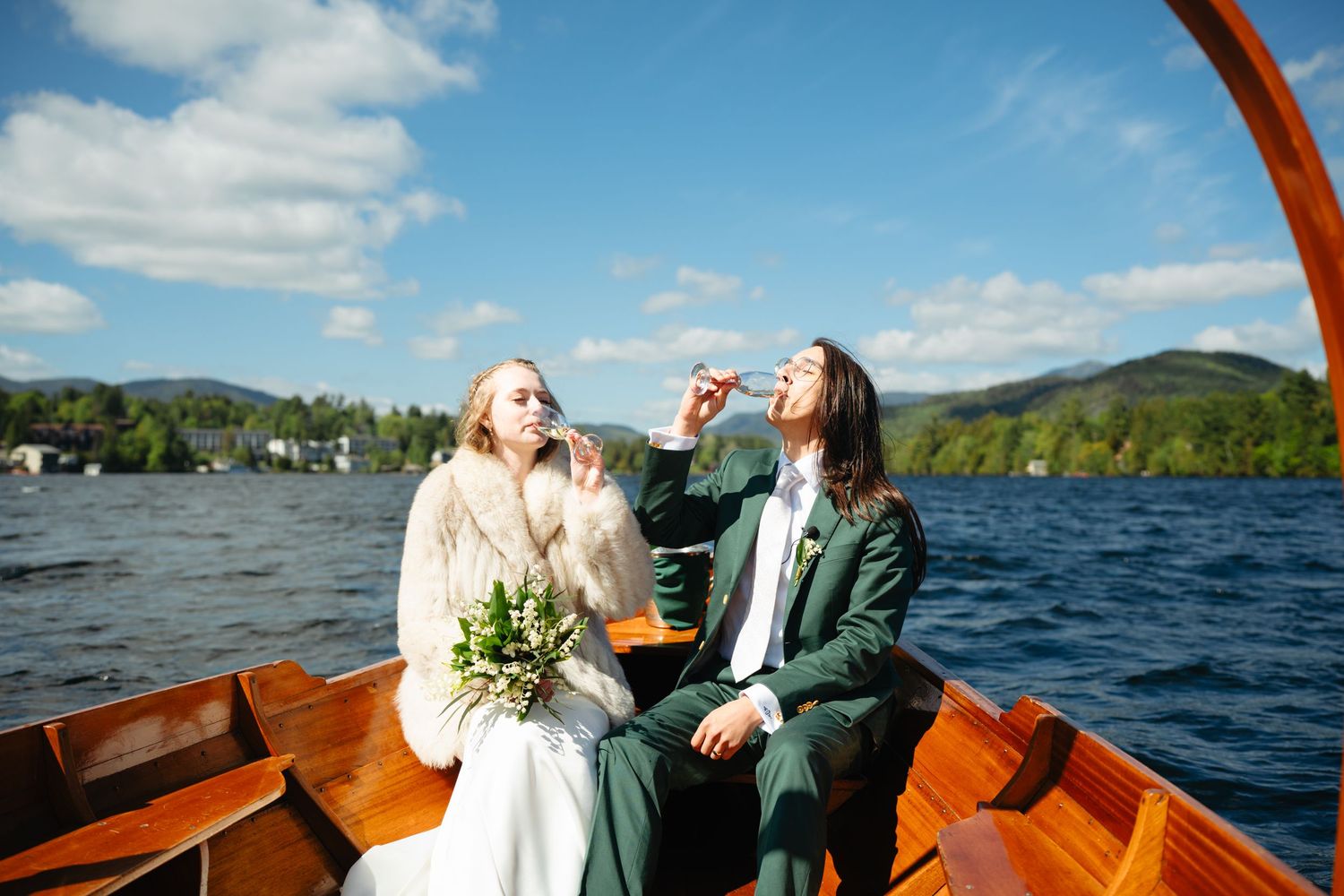 Bride and groom celebrating on a wooden boat on a lake, blowing bubbles under a bright blue sky.