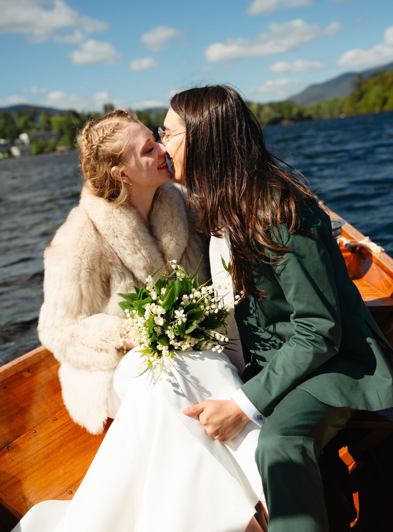 Two brides sharing a kiss on a wooden boat with a scenic mountain lake in the background.