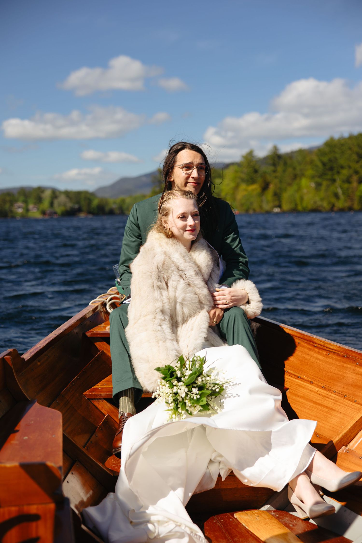 Bride and groom sit closely together in a wooden rowboat on a scenic lake on their wedding day.