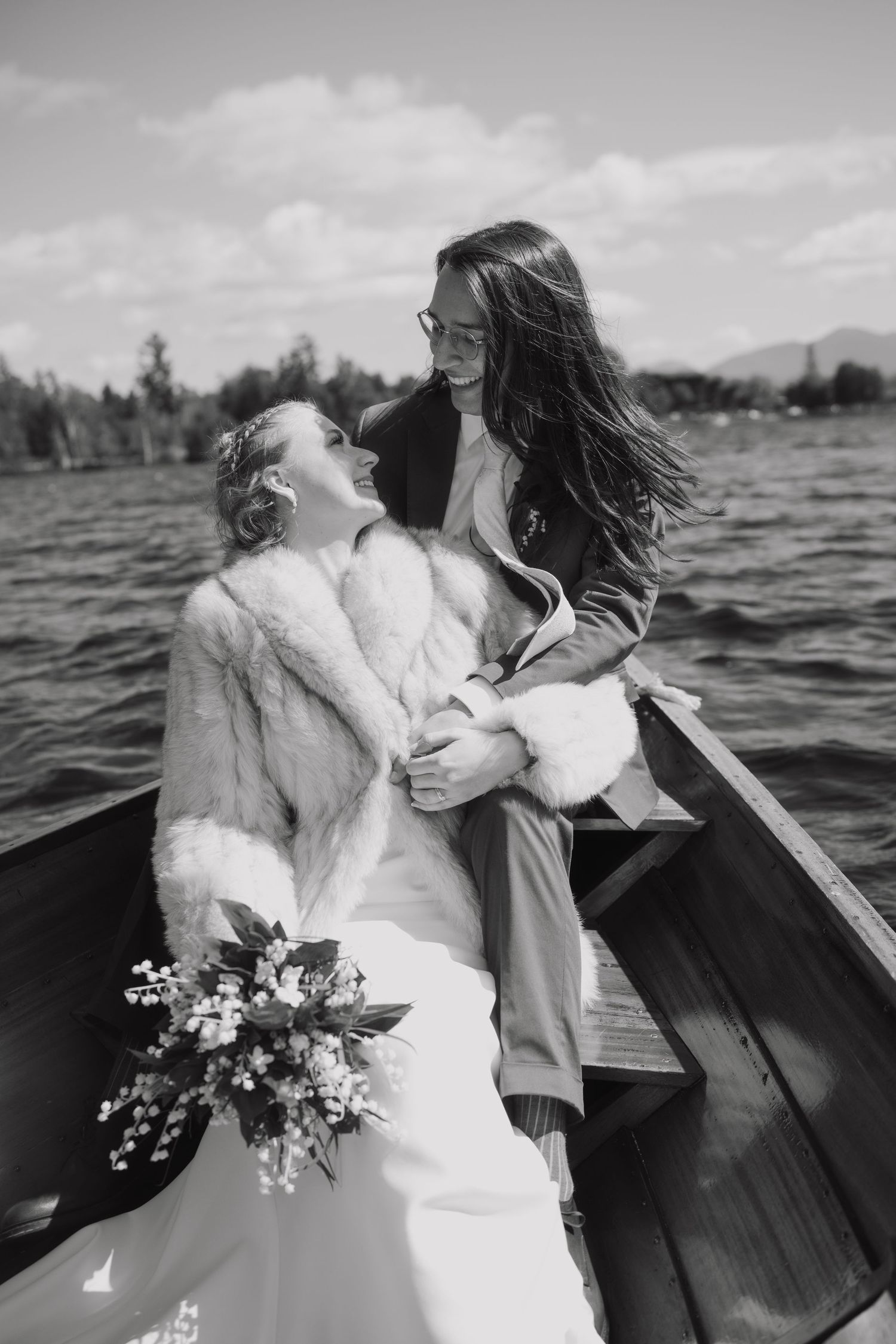 Black and white photo of two brides embracing in a wooden boat on a lake, one holding a white floral bouquet.