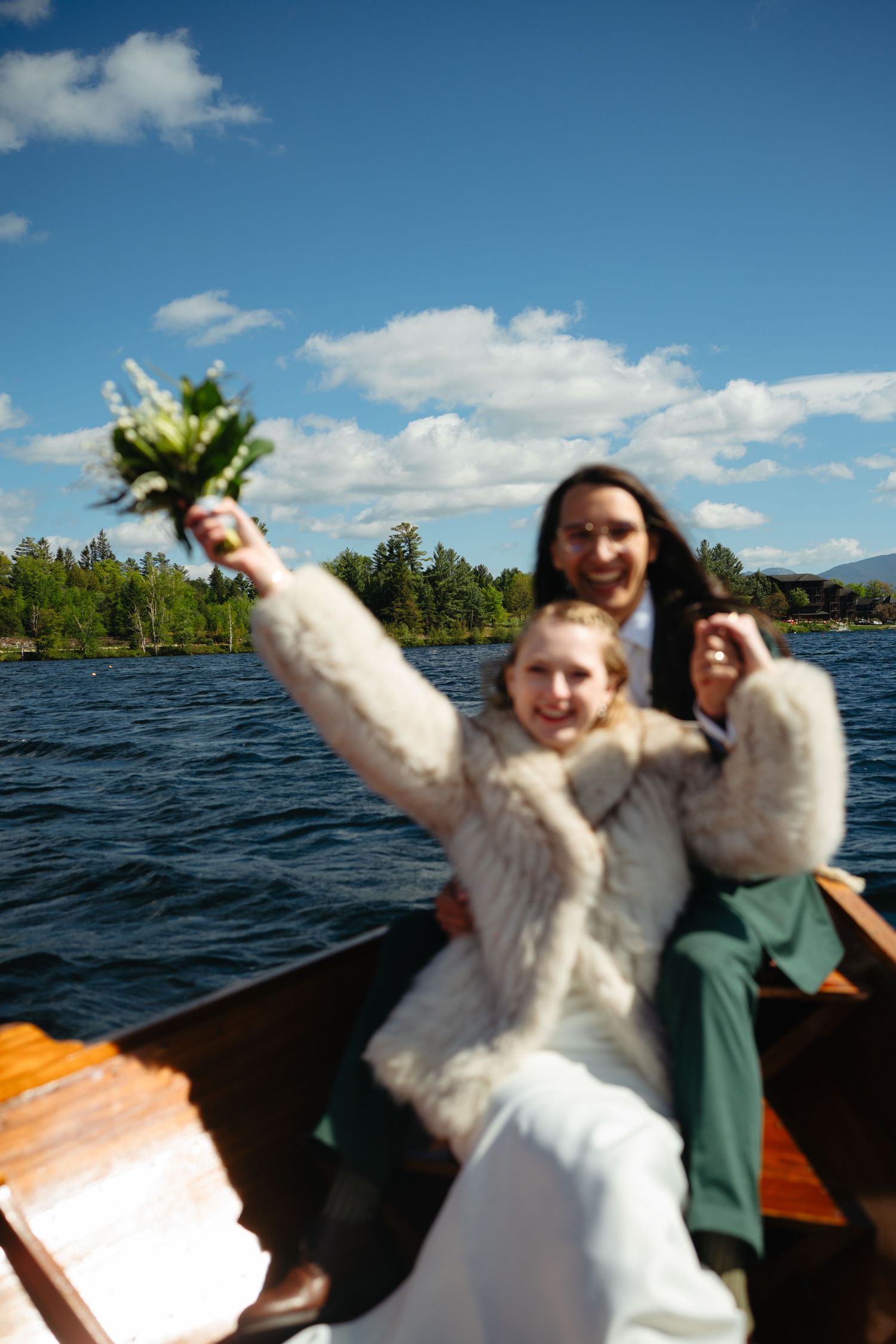 Joyful bride in white gown and fur wrap raises bouquet while riding a wooden boat on a scenic lake.