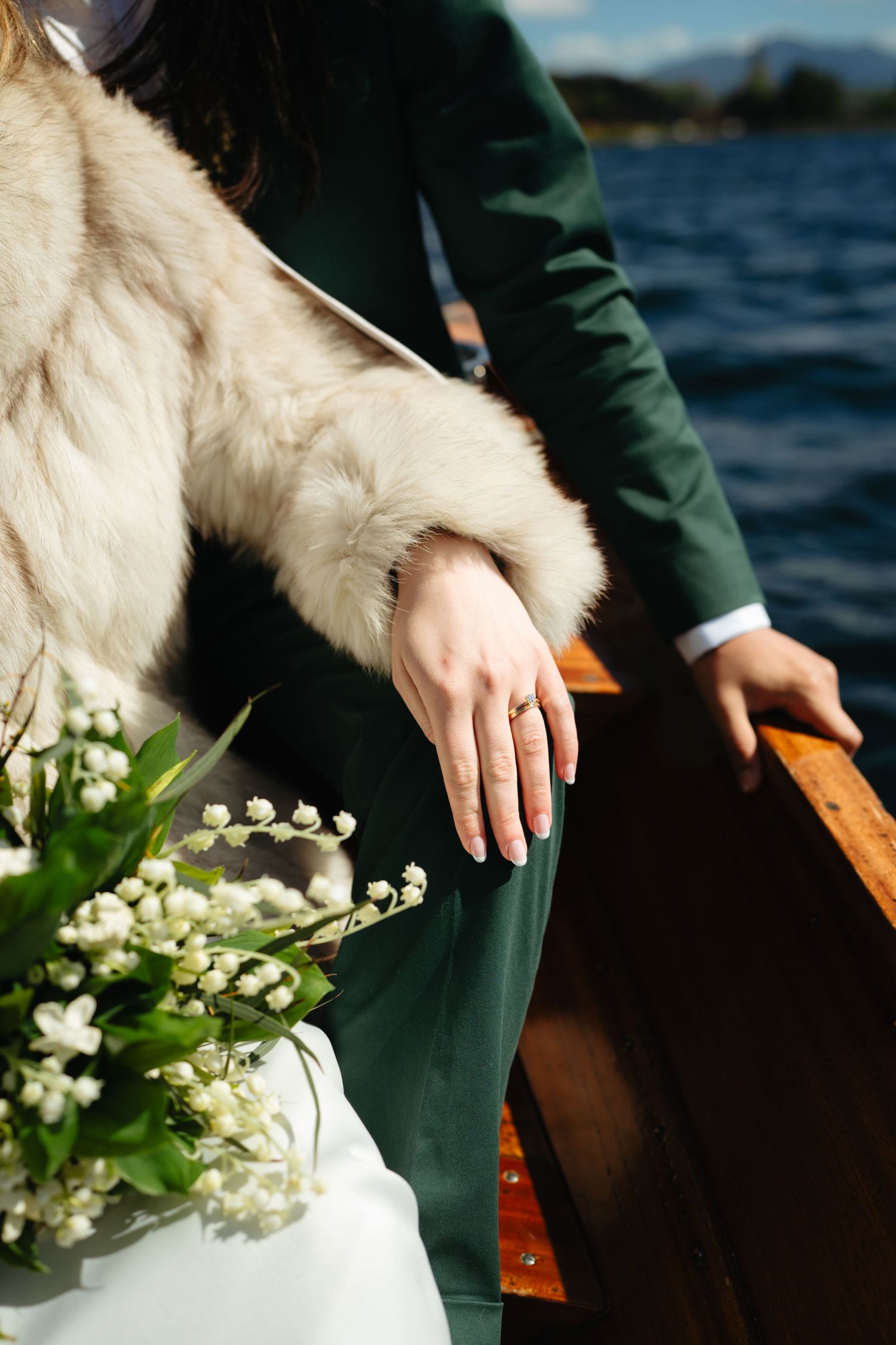 Close-up of bride's hand with wedding ring resting on wooden boat rail holding white floral bouquet.