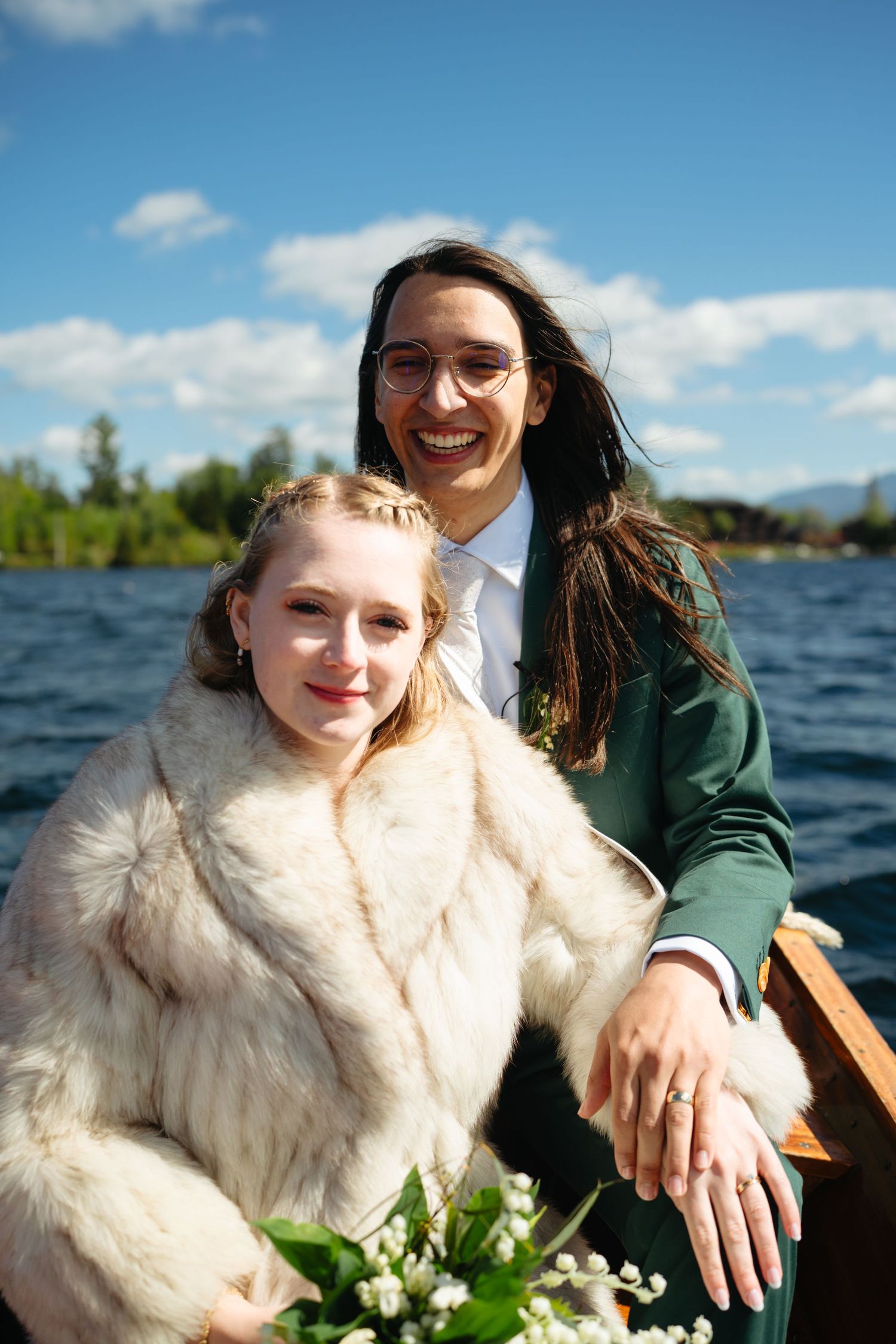 Smiling newlyweds pose on wooden boat with lake and blue sky in background.