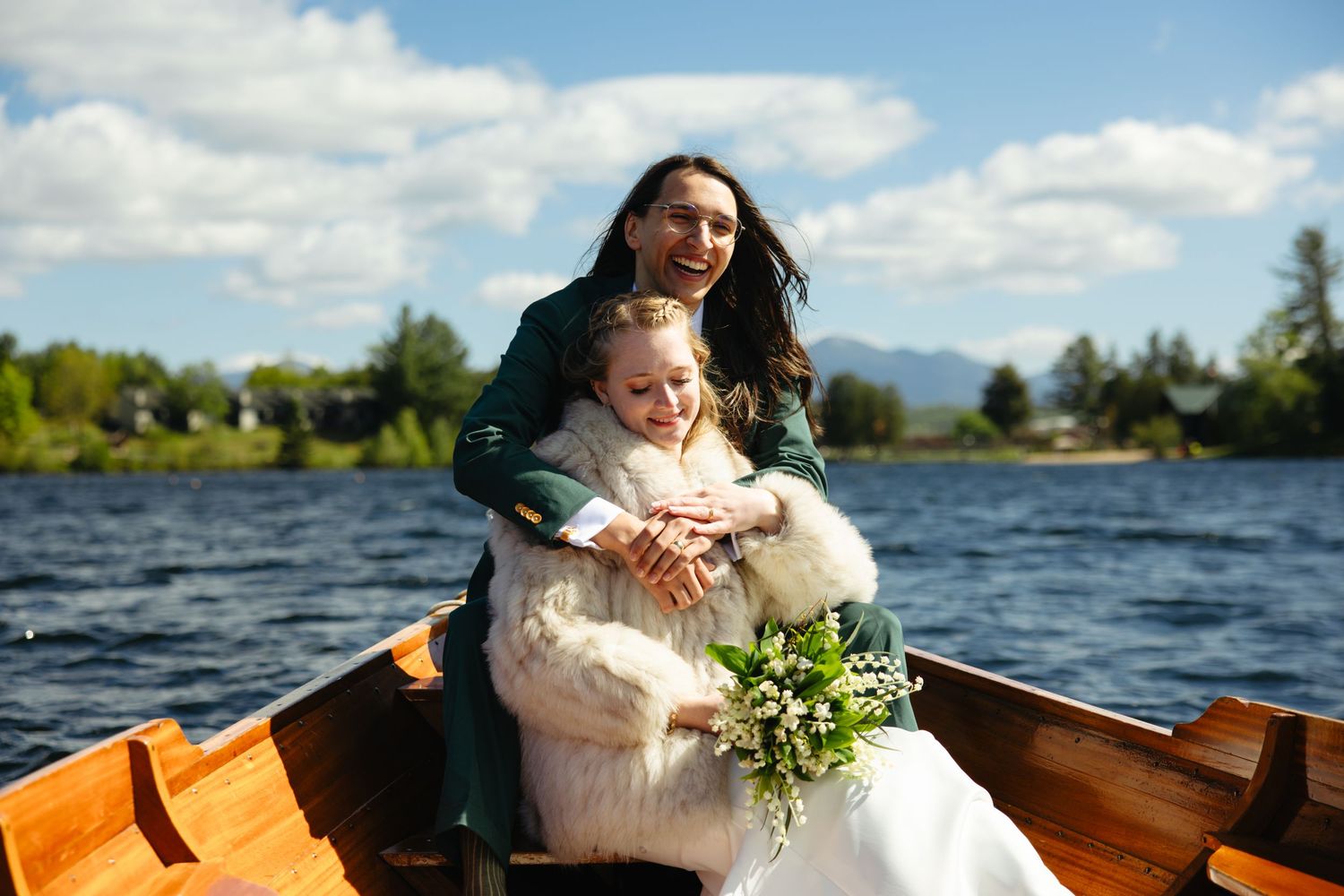 Bride and groom embrace in wooden rowboat on scenic lake under blue sky with mountains.