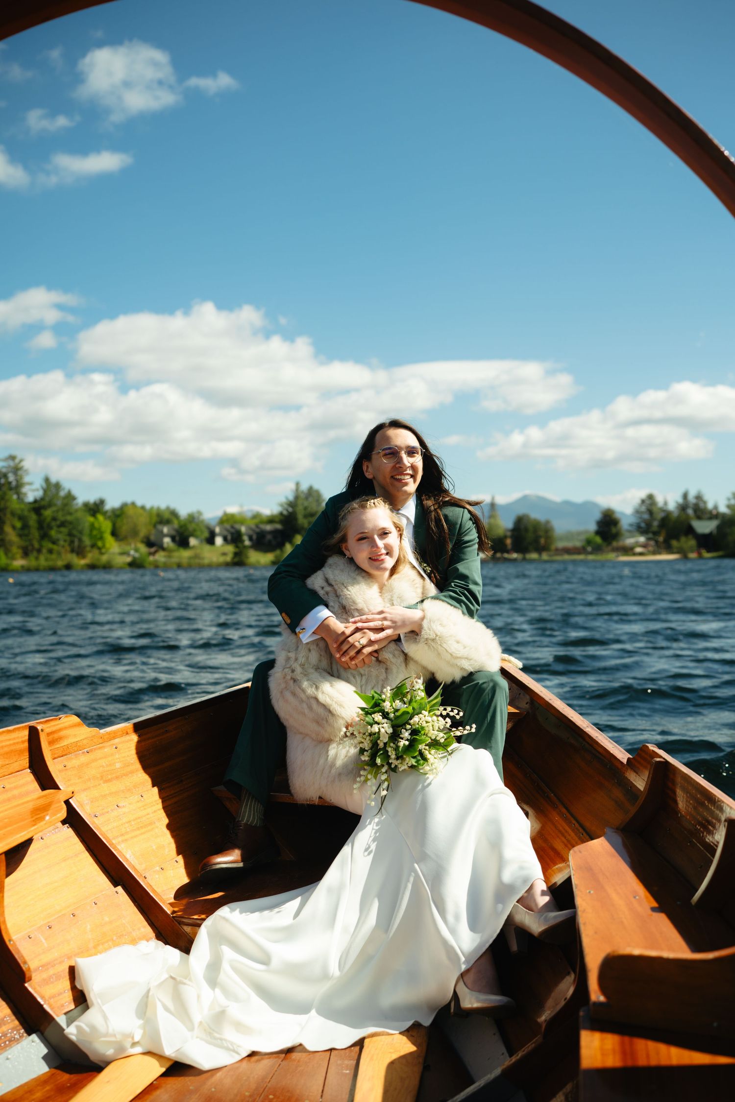 Wedding couple sitting in wooden rowboat on lake, bride holding white bouquet under sunny sky.