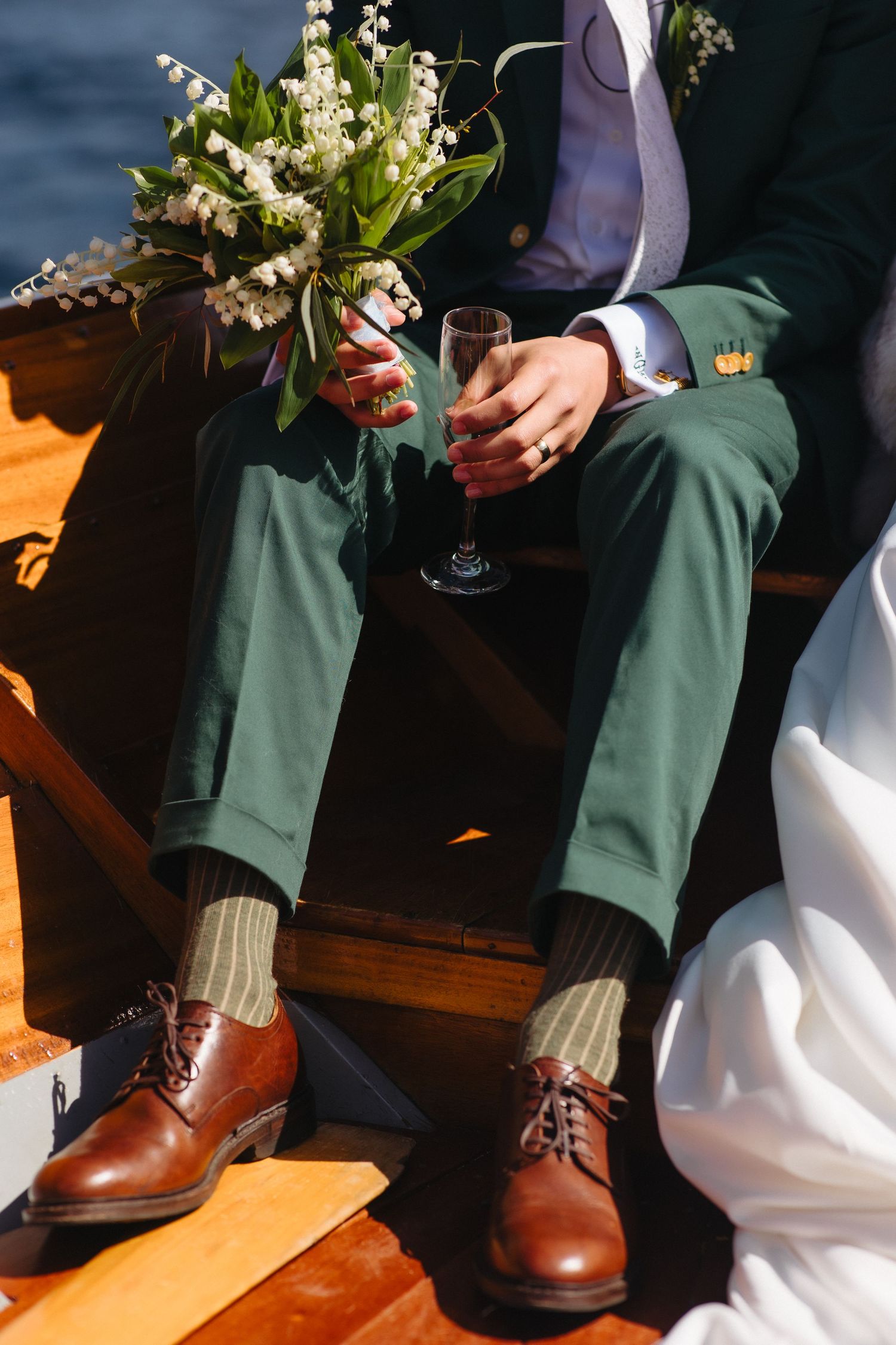 Groom in green suit holds champagne glass and bouquet while seated on wooden boat.