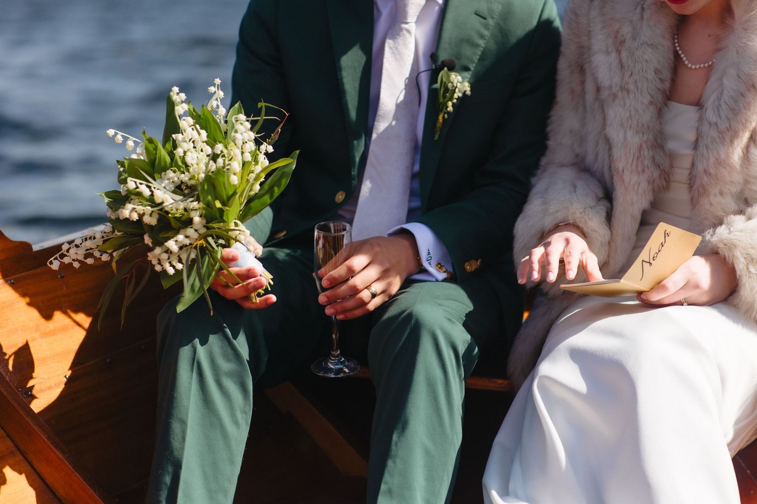 Newlyweds sit together on wooden boat holding champagne glasses and lily of valley bouquet.