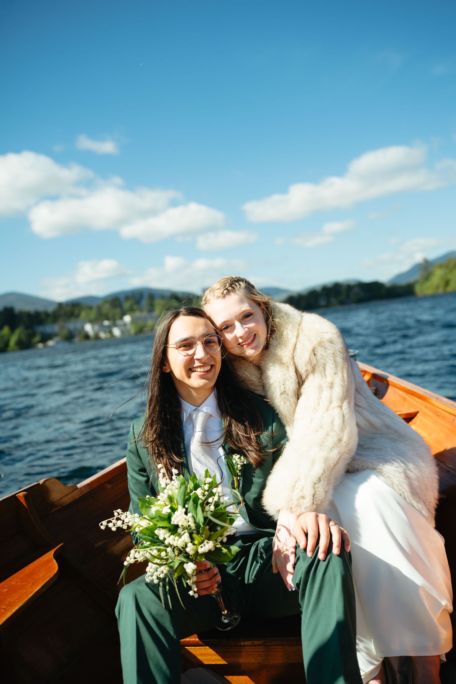 Newlyweds sitting close together in wooden boat on lake, bride leaning on groom holding bouquet.