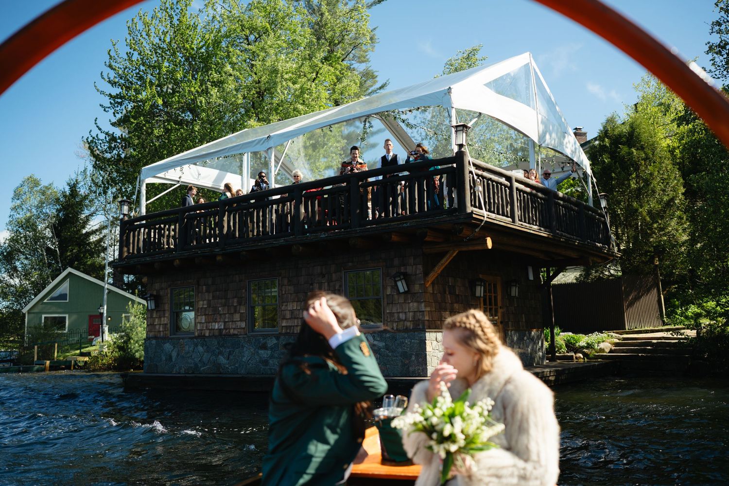 Bride and groom on a boat near a waterfront venue with guests watching from a wooden deck above.
