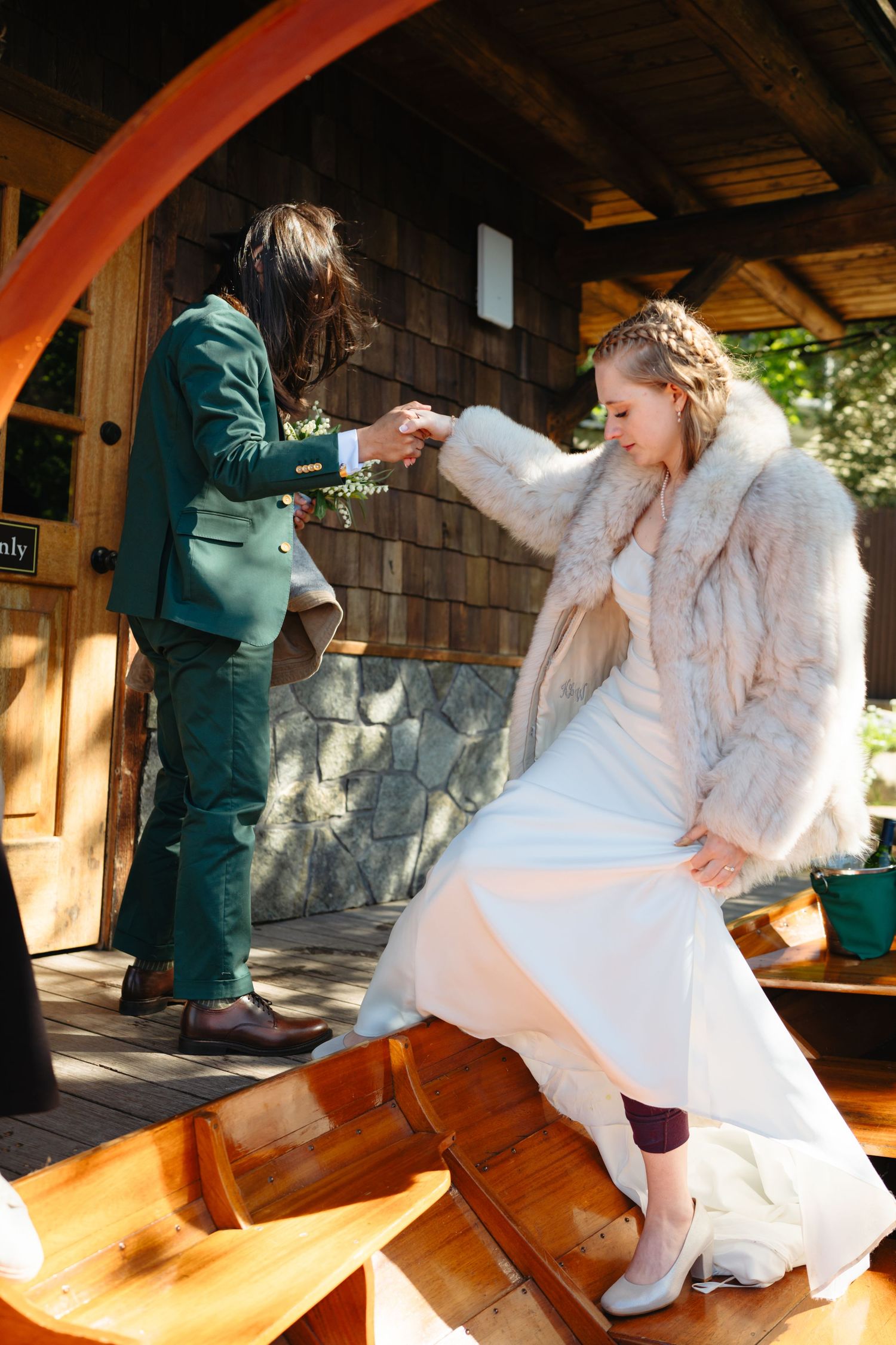 Groom in green suit helps bride in fur coat step off wooden boat at dock.