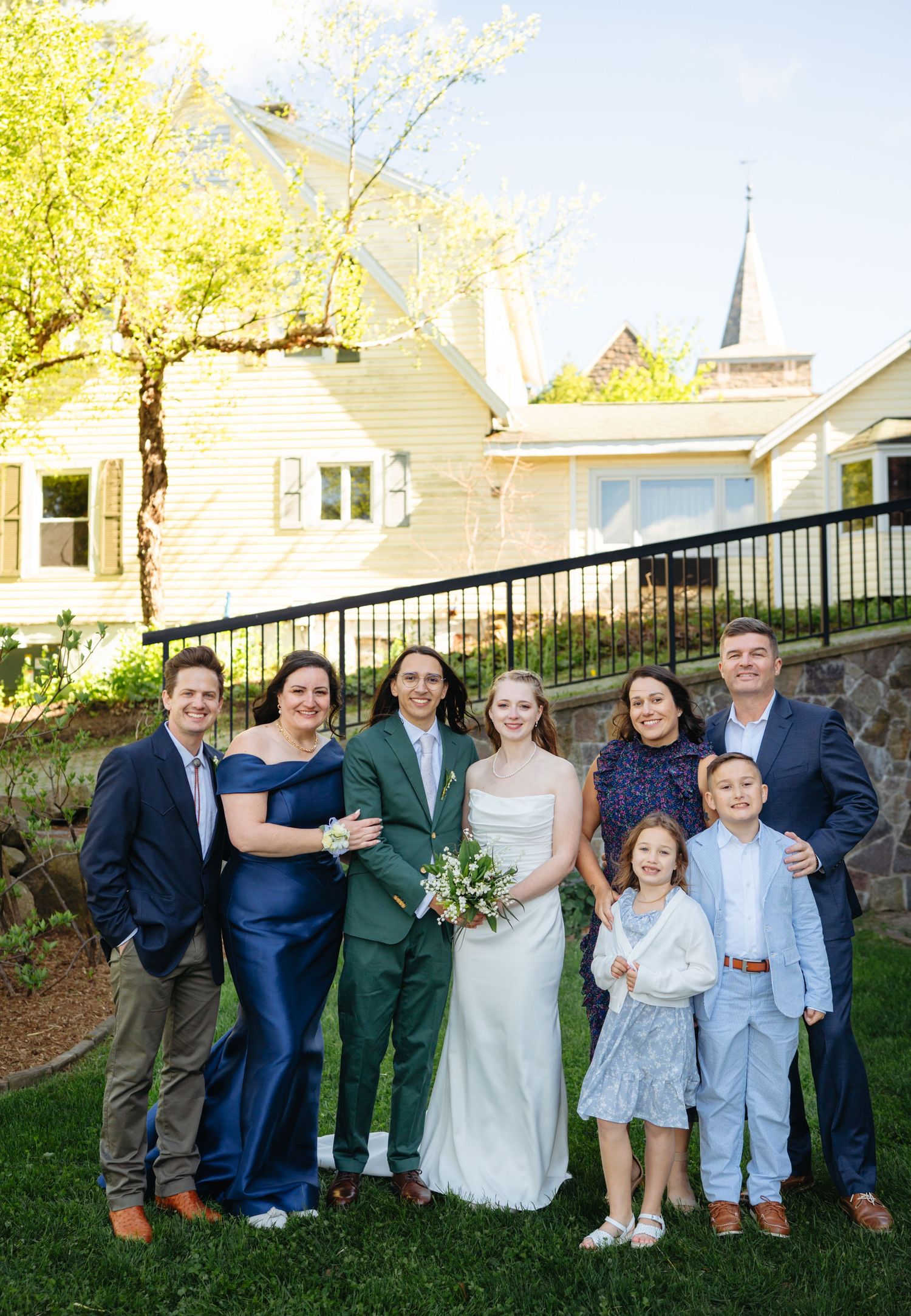 Wedding couple in green and white attire posing with family group outdoors near historic church building.