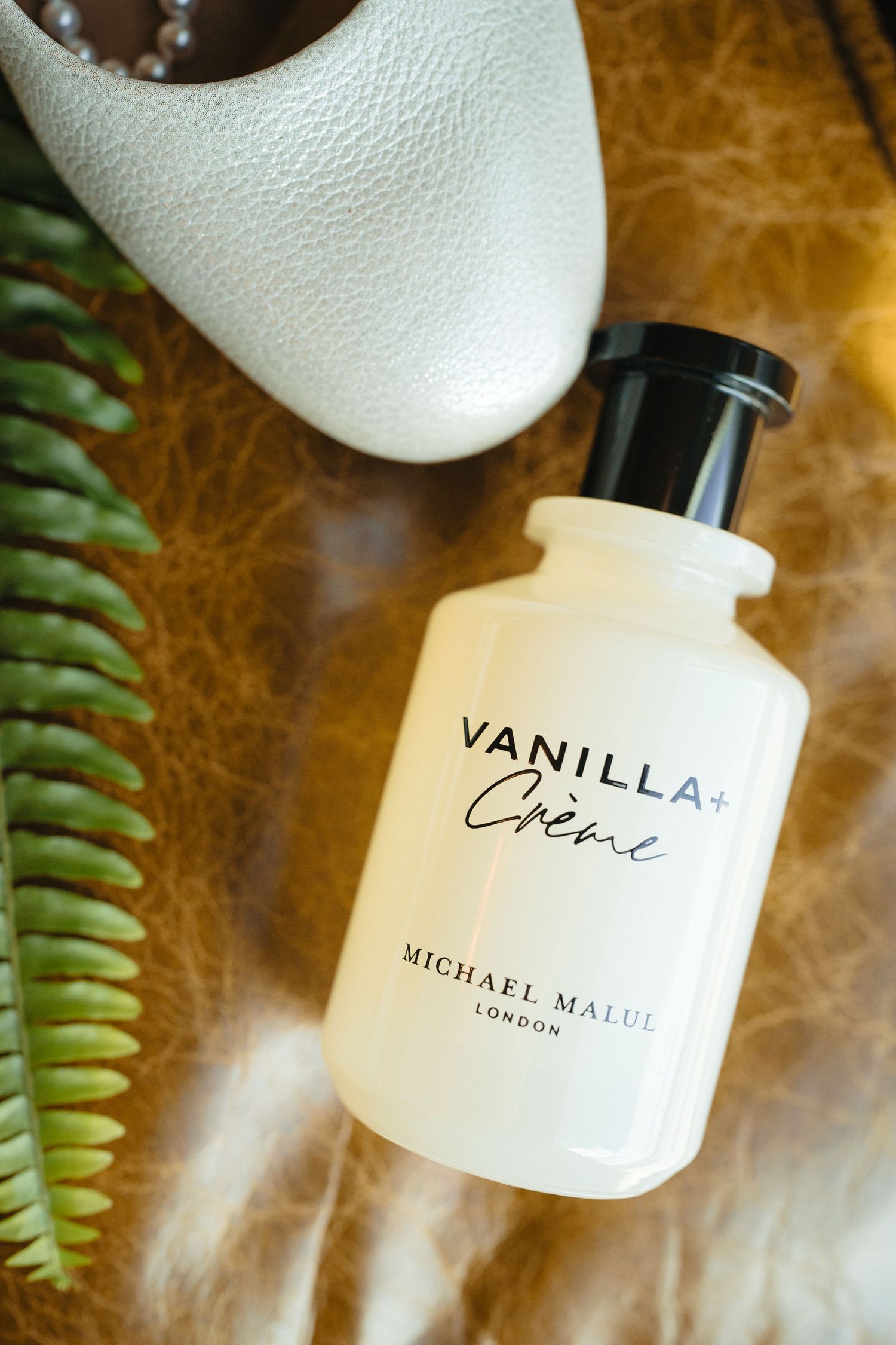 Vanilla Creme perfume by Michael Malul London displayed with white heels and fern leaves on brown leather.