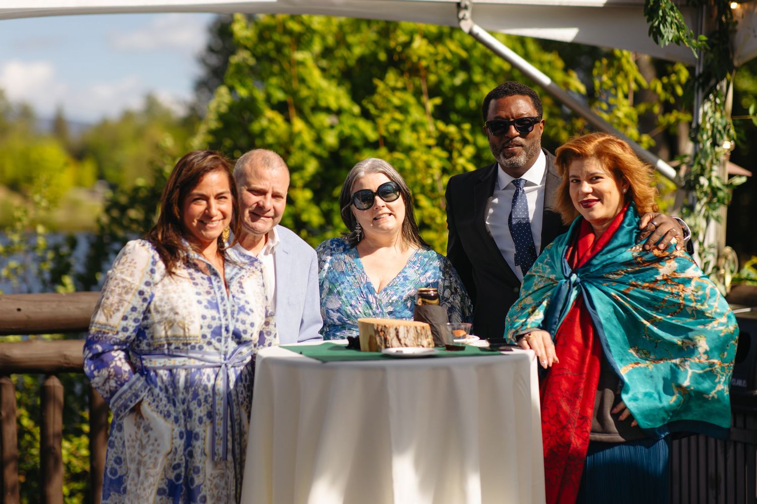 Five elegantly dressed wedding guests pose together outdoors at a reception table with lush greenery behind them.