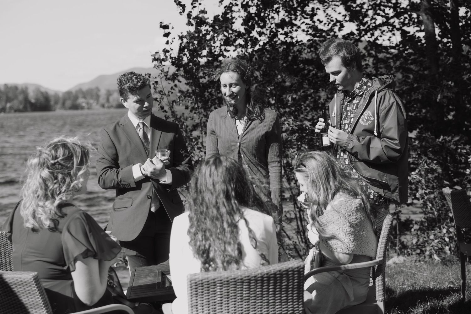 Black and white photo of wedding guests gathered outdoors under trees in mountain setting.
