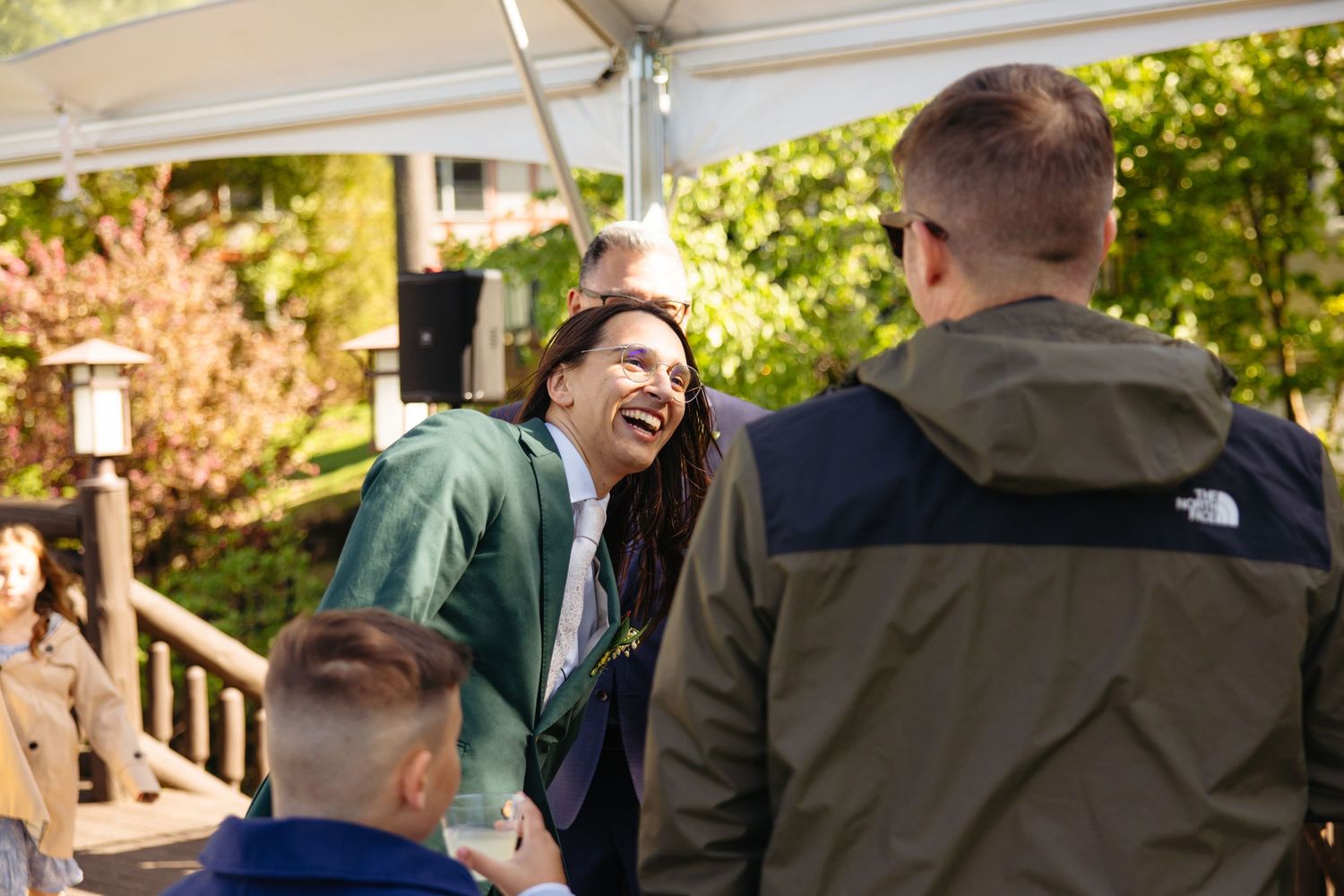 A woman in a green jacket laughs joyfully while chatting with guests at an outdoor wedding celebration.