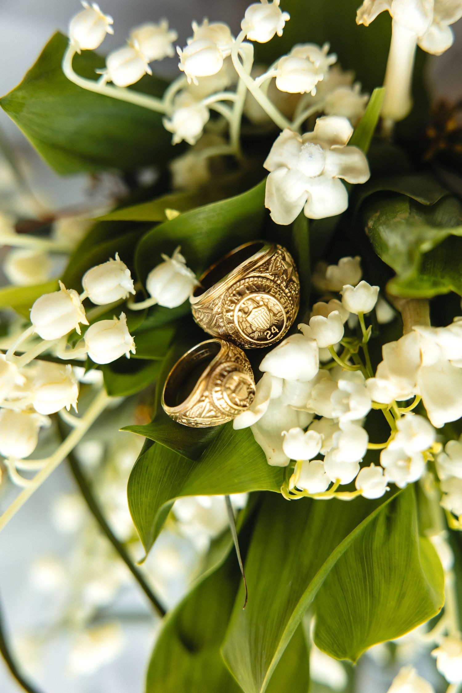 Two gold signet rings nestled among white lily of the valley flowers and green leaves in a bridal bouquet.