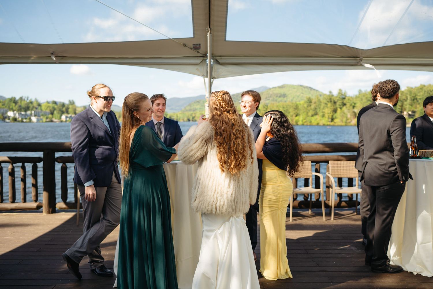 Wedding guests mingle on a lakeside deck under a white canopy tent on a sunny day.