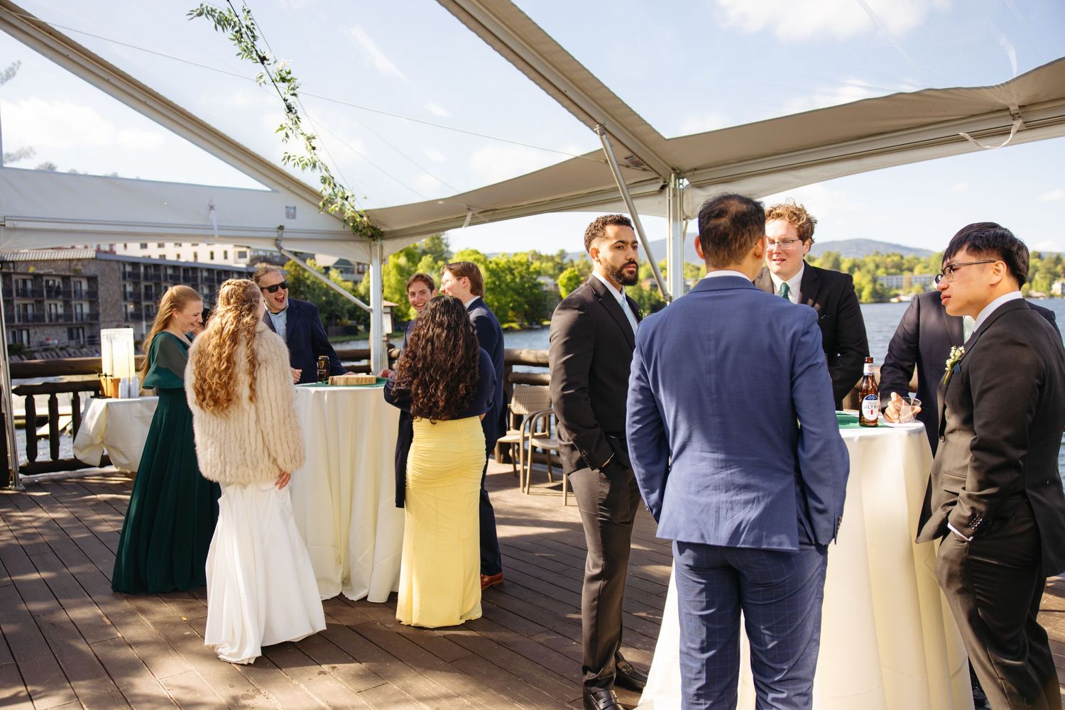 Wedding guests in formal attire socialize at an outdoor waterfront cocktail hour under a tent.