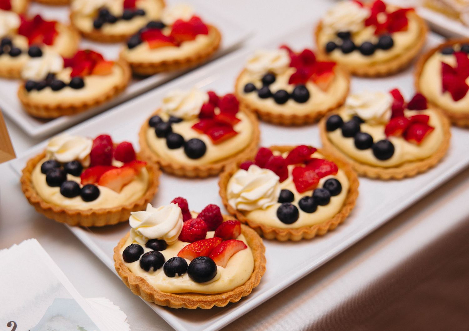 Assorted fruit tarts with blueberries, raspberries, and cream on a white platter at a dessert display.