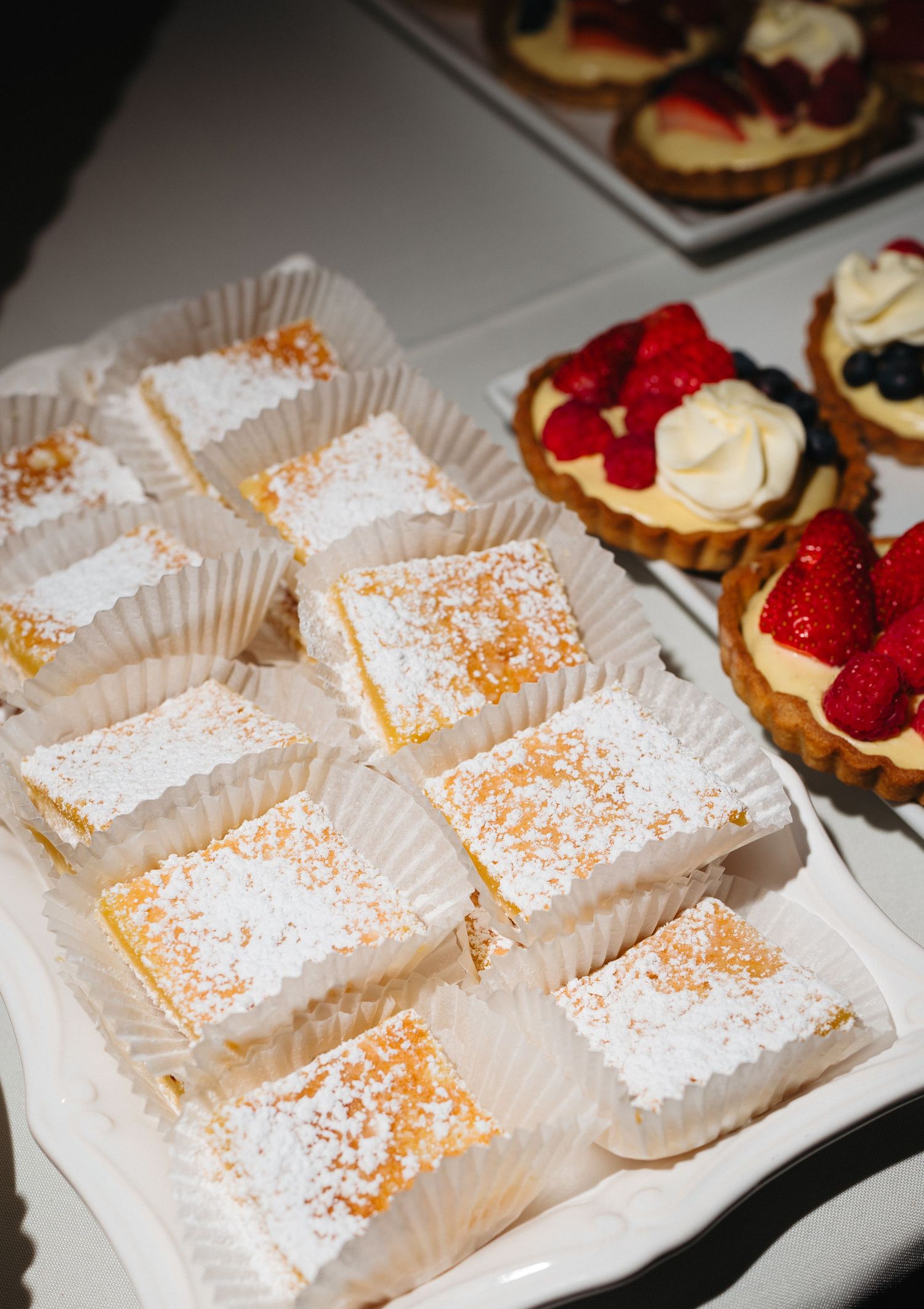 Powdered sugar lemon bars and fresh berry tarts displayed on white platters at wedding dessert table.