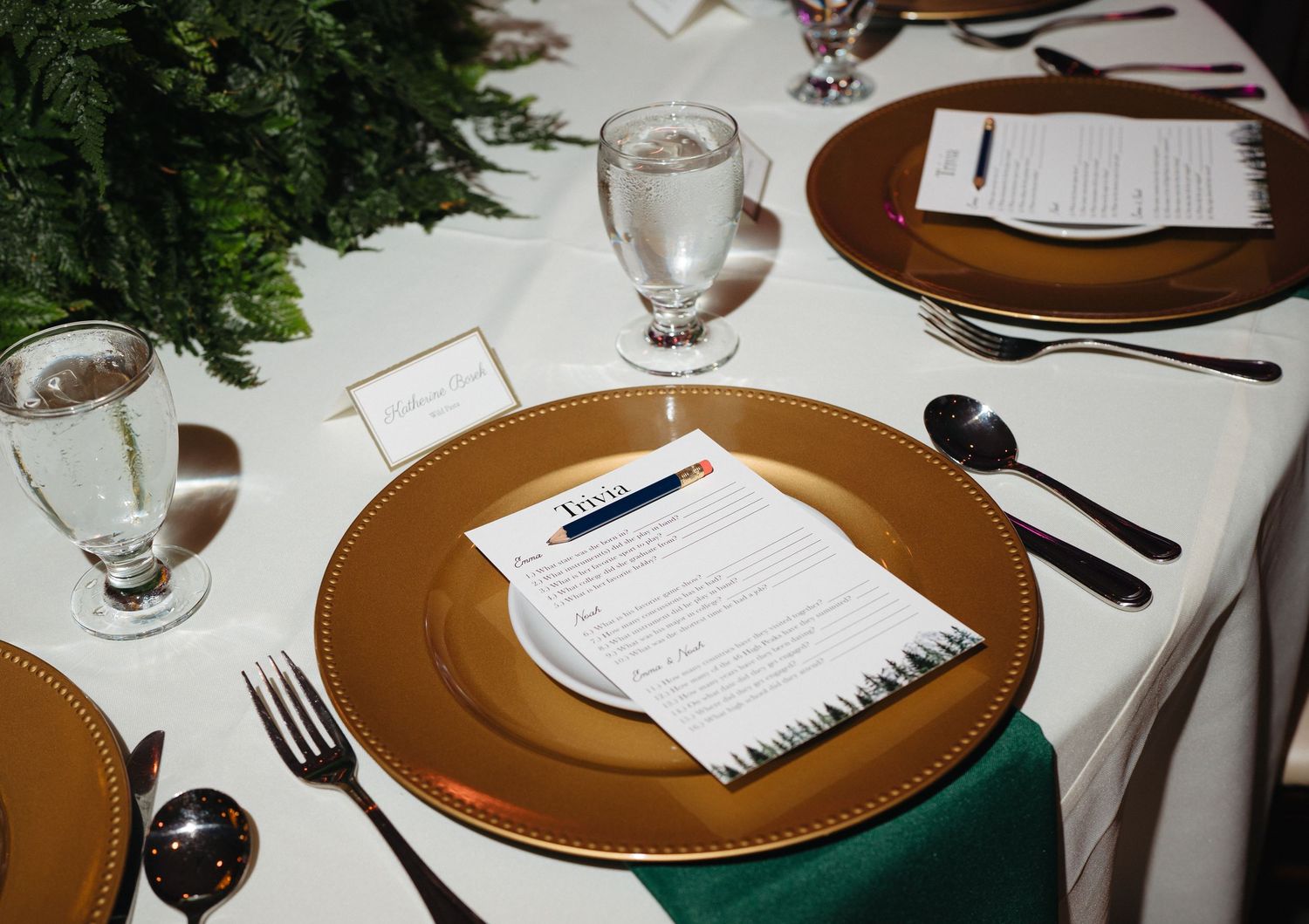 Elegant wedding place setting with gold charger plate, menu card, and green napkin on white tablecloth.