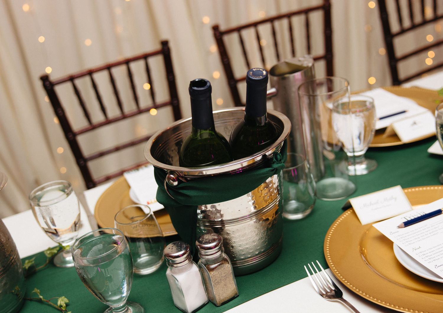 Elegant wedding reception table with green tablecloth, gold charger plates, wine bottles in ice bucket, and glassware.